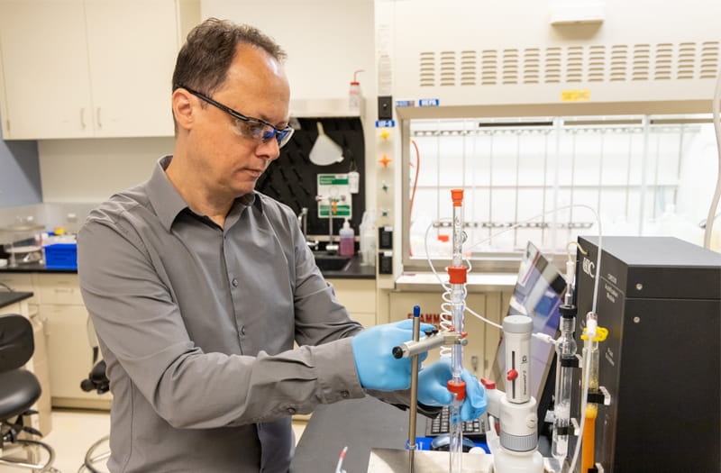 Radu Custelcean, wearing a gray shirt and safety glasses, manipulates an acidification module and other chemistry equipment in a laboratory.