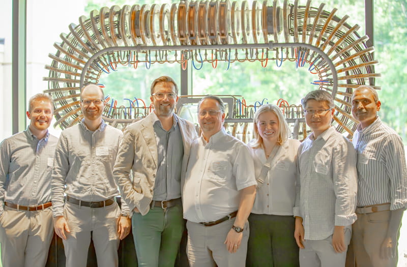 Seven people in business casual clothes standing in front of a model of a stellarator consisting of a metal oval with colorful wires and copper-colored coils.