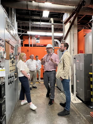 Five people on a facility tour stand near banks of grey control panels.