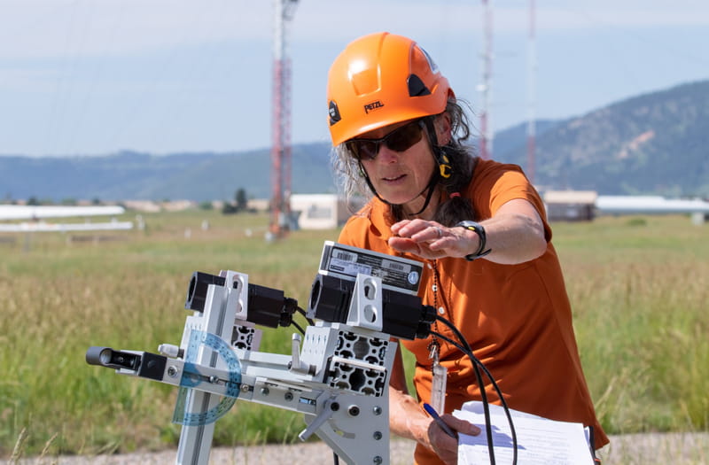 Wearing an orange shirt and orange hard hat and holding sheets of paper and a pen, a scientist examines a complex array of metal and plastic components. In the background, grass extends to a line of tall hills.
