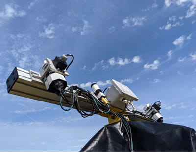 Posed against a blue sky studded with small windswept clouds, a horizontal metal bar with wires and multiple small components emerges from a sheet of heavy black fabric.