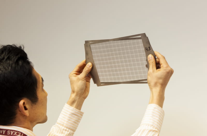 A view over the shoulder of a man holding two overlapping metal sheets over his eye level. The silver metal sheets each have a narrow frame that around a rectangular area perforated with many small holes. The holes in the two sheets line up to create a moire pattern.
