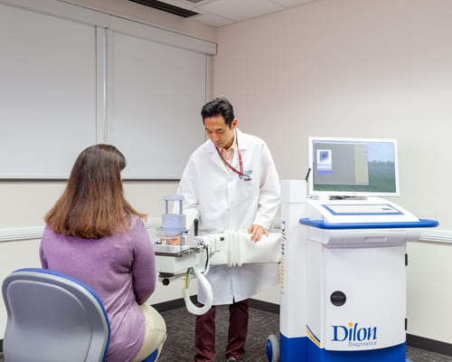 A woman sits in a room, facing away from the camera to look toward the arm extending from a white roughly cylindrical piece of equipment. The top of the cylinder holds a computer screen and keyboard. A man adjusts the arm extending from the cylinder.