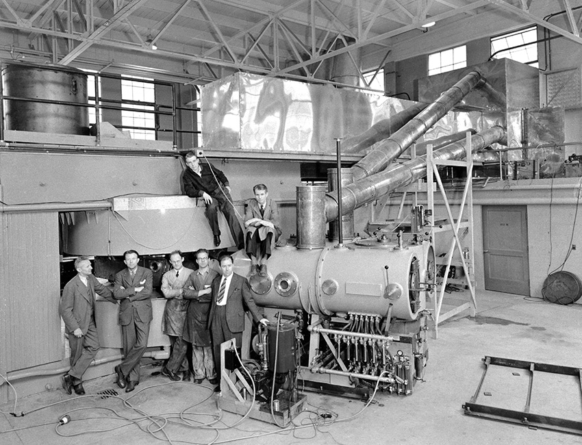A black and white photo of 7 men in suits posing in front of large machinery from decades ago