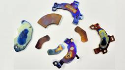 A photo of four voice coil magnets taken from a desktop or laptop computer and their brackets (which have mounting holes).  These magnets are each about 10.5 – 15 grams, 1 - 1.25 inches long.  These ones are colorful, because they have been overheated in the process of separation and demagnetization. Photo supplied by the Ames Laboratory.