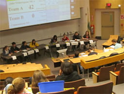 An audience looks on in an auditorium as students sit at the front of the room at tables competing in the Science Bowl regionals