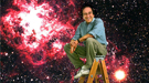 Scientist Saul Perlmutter sitting atop a ladder with a large image of the universe in the background