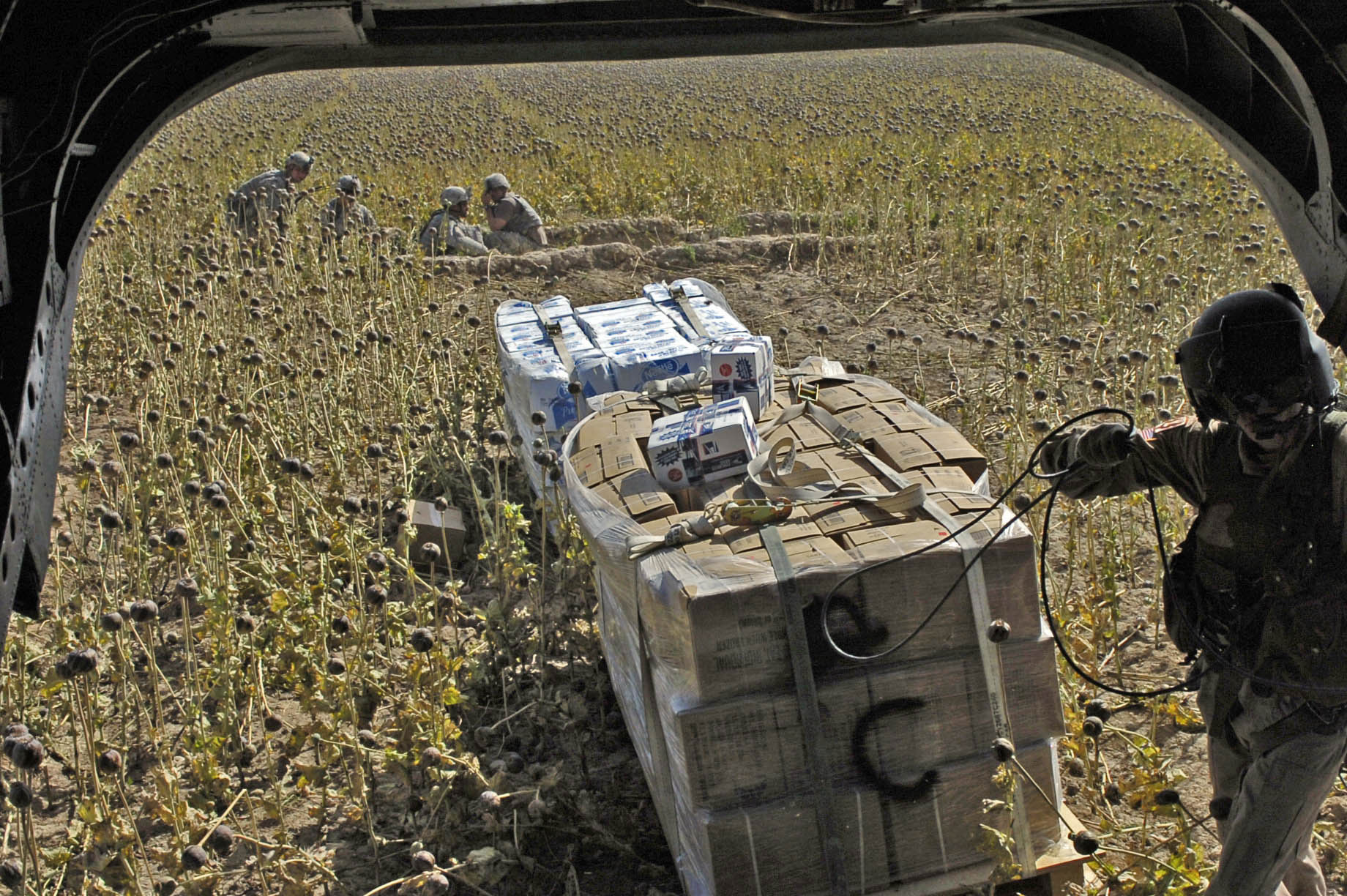 Boxes of water being offloaded from the back of a military helicopter into a field with 4 soldiers waiting to unload