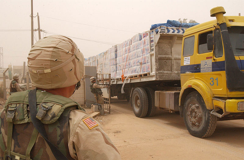 A US soldier in the foreground looking over a large cargo truck carrying cases of water