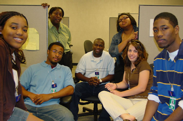 Group of seven casually dressed people in a work environment posing for a photo near cubicles