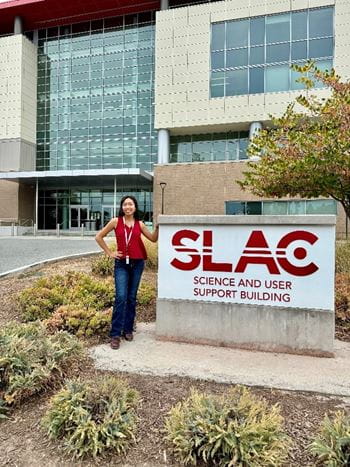 Megan Rogers standing beside the SLAC Science and User Support Building sign near the entrance to SLAC.