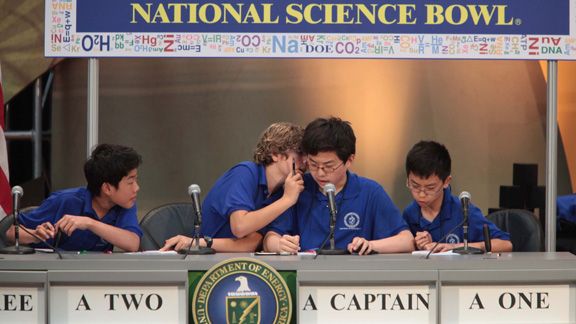 Four Middle School boys in blue shirts sitting a table during a match with one whispering in another's ear